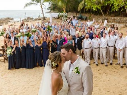 A bride and groom kissing on the beach while their wedding guests cheer behind them